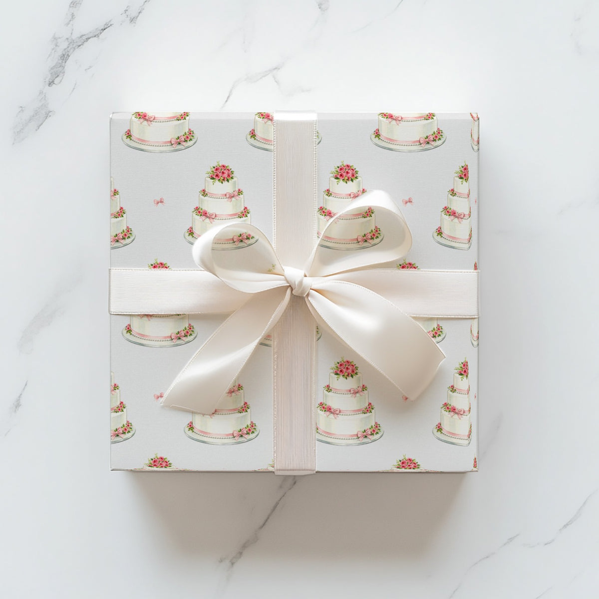 Square gift wrapped in minimalist wedding cake wrapping paper with an ivory satin ribbon and bow on a white marble surface