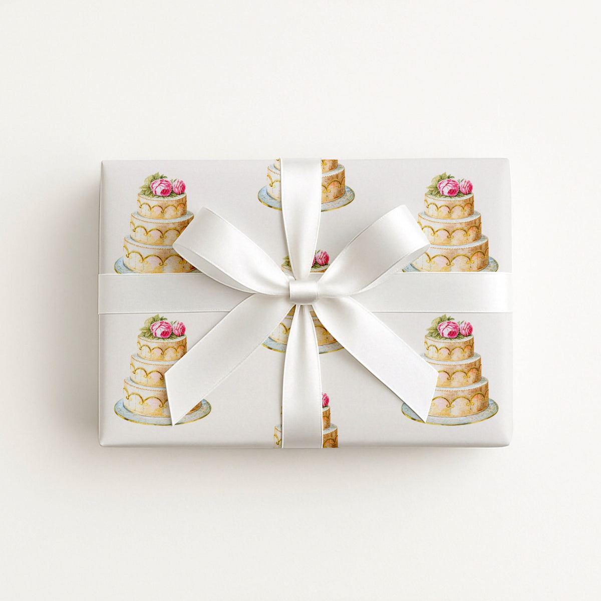 Flat lay of a gift wrapped in elegant wedding cake wrapping paper with tiered cakes and pink roses, tied with a white satin ribbon and bow