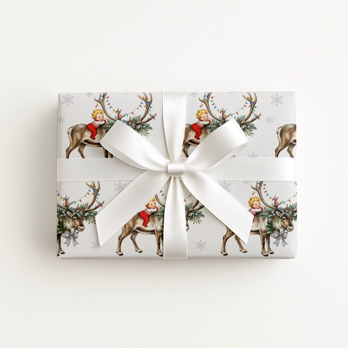 A rectangular gift box wrapped in ivory wrapping paper with a vintage baby and reindeer print, tied with a large white satin ribbon bow, on a clean white background.