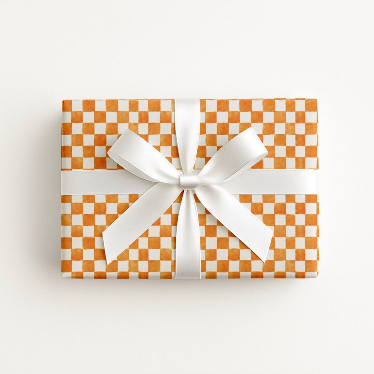 A rectangular gift box wrapped in orange and white checkered paper, tied with a wide white satin ribbon and bow, photographed from above on a clean white background.