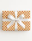 A rectangular gift box wrapped in orange and white checkered paper, tied with a wide white satin ribbon and bow, photographed from above on a clean white background.