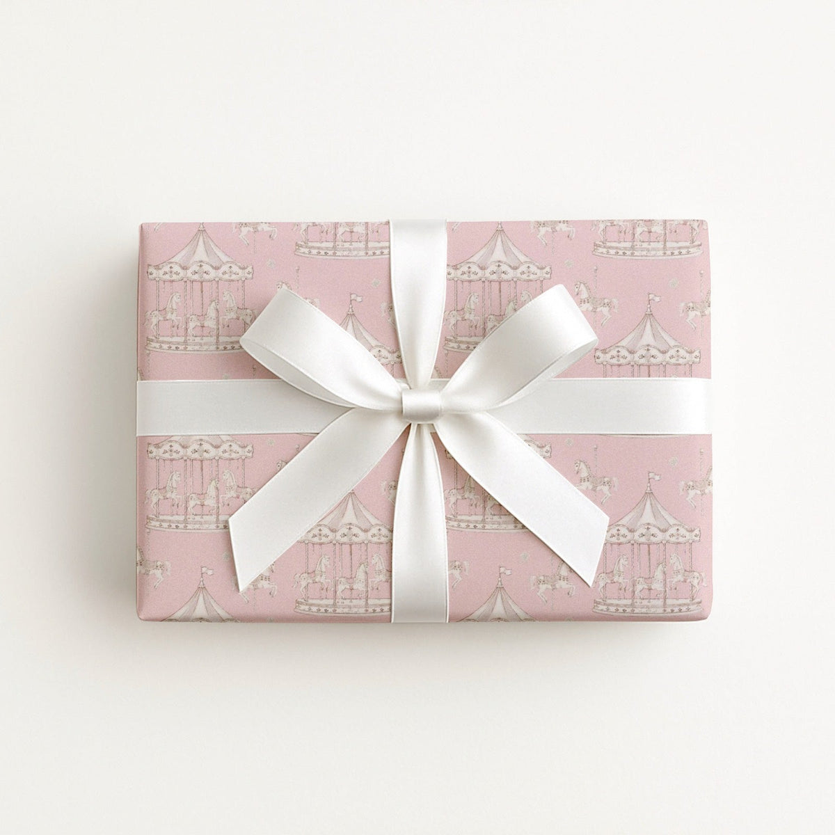 A square gift box wrapped in light pink paper with a french carousel pattern, tied with a wide white satin ribbon.