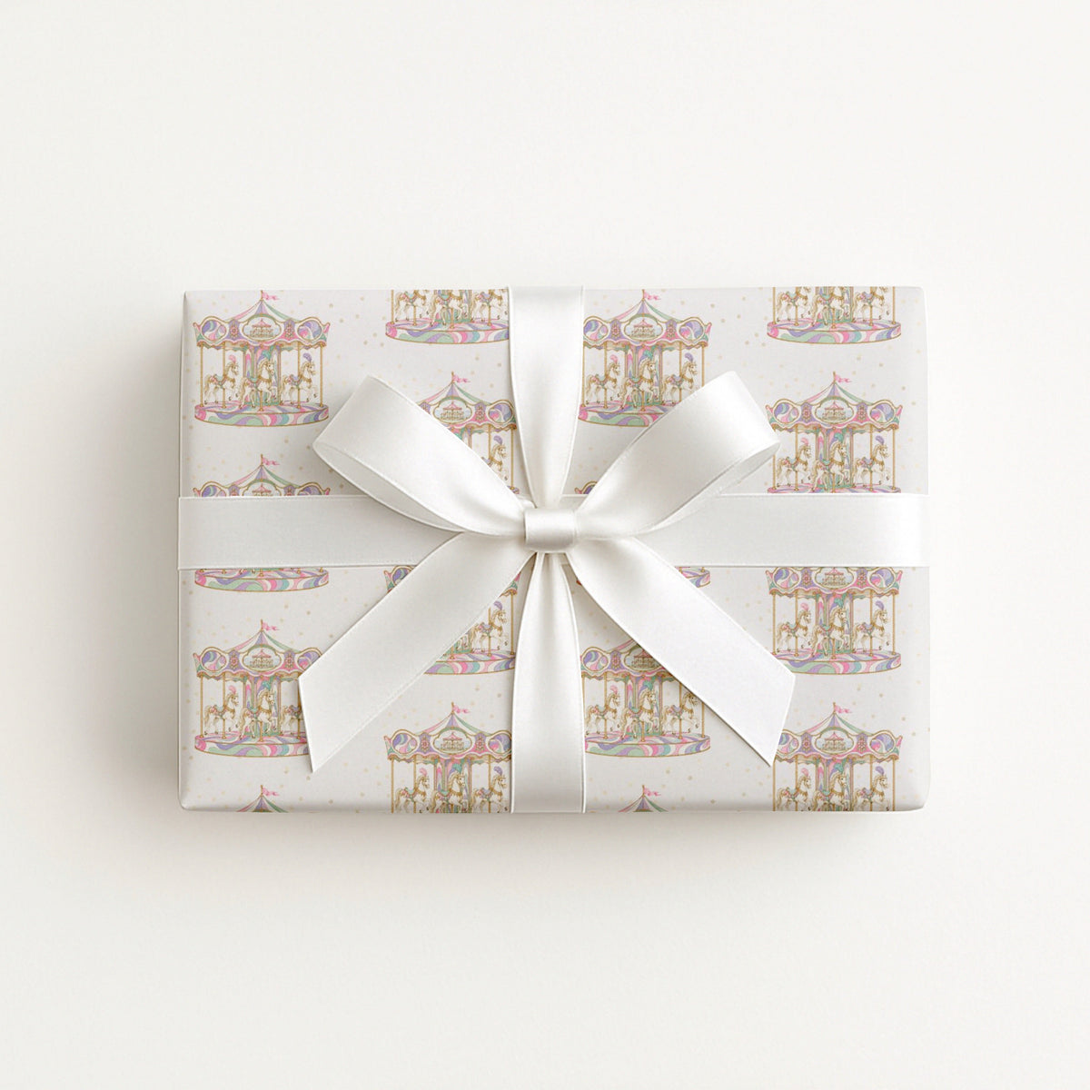 A rectangular gift box wrapped in white paper with a repeating pastel carousel pattern, tied with a wide white satin ribbon and bow, photographed from above on a plain white background.