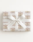A rectangular gift box wrapped in white paper with a repeating pastel carousel pattern, tied with a wide white satin ribbon and bow, photographed from above on a plain white background.