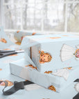 Light blue wrapping paper with a illustrations of a vintage bride, shown wrapped around gift boxes on a table with scissors and ribbon in a bright indoor setting.