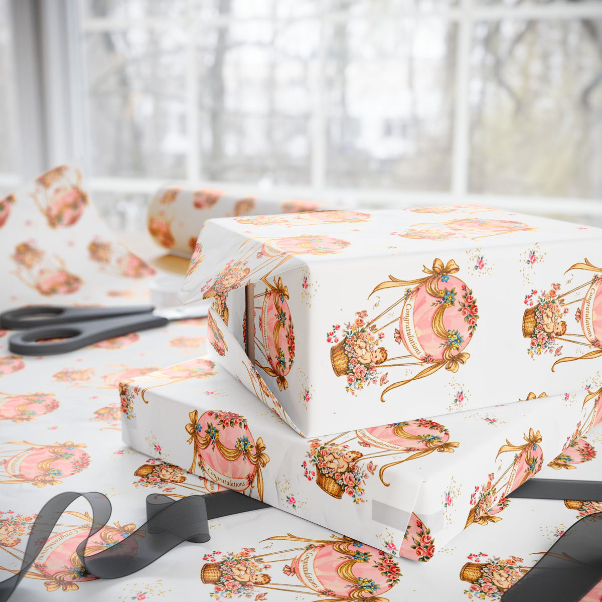 Three gift boxes wrapped in cream baby shower wrapping paper with a pink and gold vintage hot air balloon pattern, styled on a table with scissors and ribbon in front of a softly blurred window.