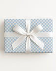 A rectangular gift box wrapped in light blue and white checkered paper, tied with a wide white satin ribbon and bow, photographed from above on a clean white background.