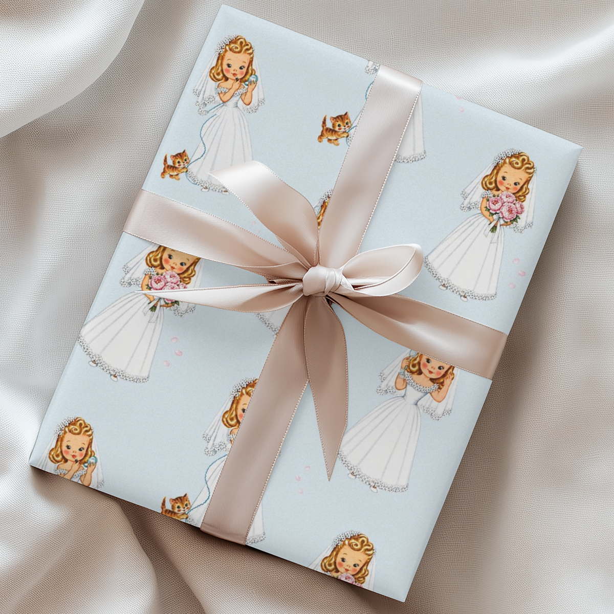 A gift wrapped in powder blue paper printed with retro brides in white dresses and a wide pink satin ribbon tied in a bow, photographed on draped satin fabric