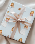 A gift wrapped in powder blue paper printed with retro brides in white dresses and a wide pink satin ribbon tied in a bow, photographed on draped satin fabric