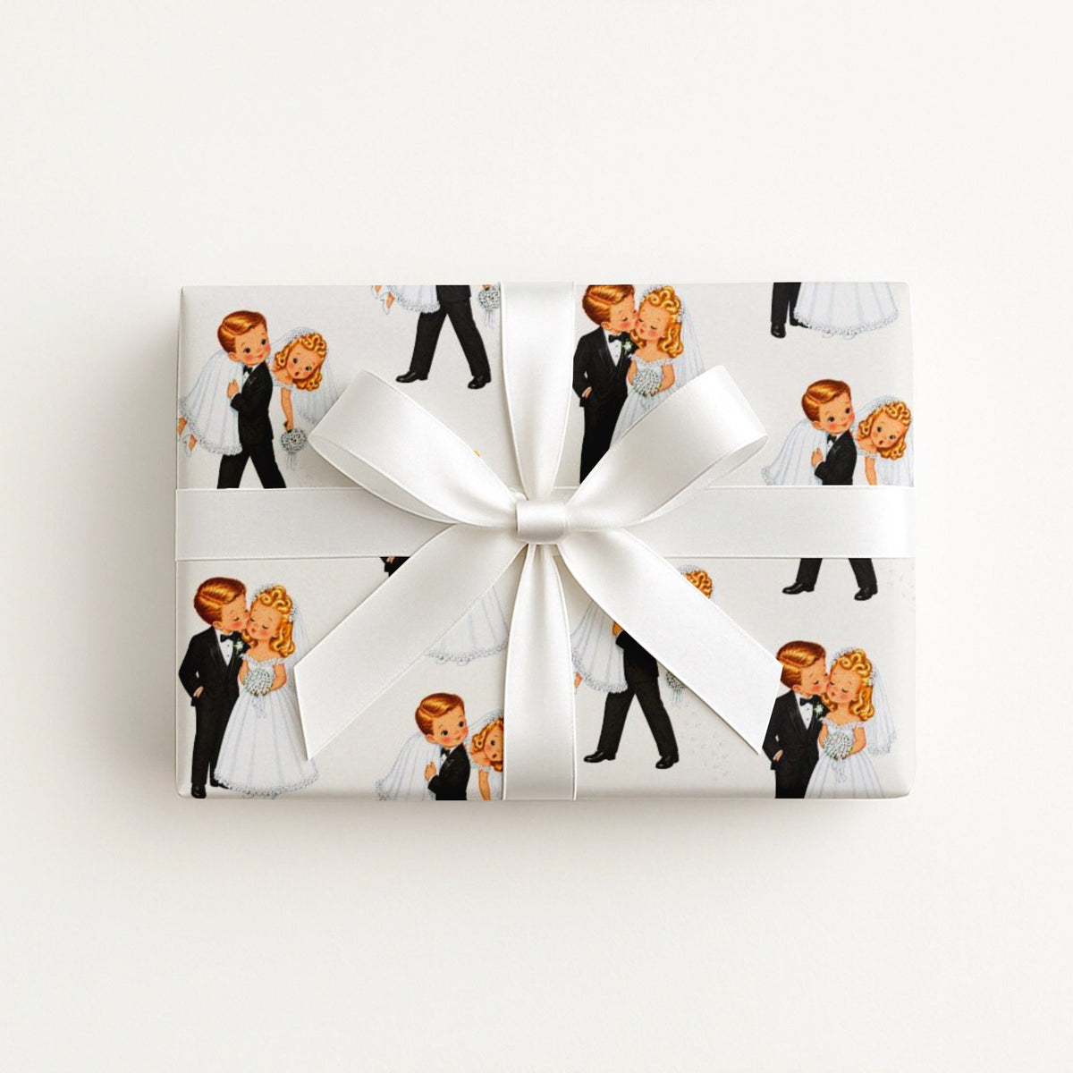 A rectangular gift box wrapped in white paper featuring a repeating pattern of vintage-style illustrated bride and groom, tied with a large white satin bow in the center, on a plain white background.
