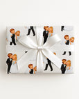 A rectangular gift box wrapped in white paper featuring a repeating pattern of vintage-style illustrated bride and groom, tied with a large white satin bow in the center, on a plain white background.