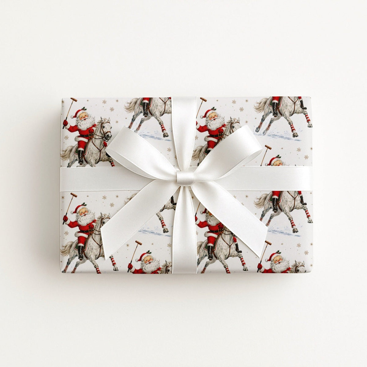 A rectangular gift box wrapped in ivory wrapping paper featuring a vintage Santa Claus riding a horse and playing polo, tied with a wide white satin ribbon and bow.