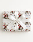A rectangular gift box wrapped in ivory wrapping paper featuring a vintage Santa Claus riding a horse and playing polo, tied with a wide white satin ribbon and bow.