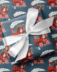 A gift box wrapped in blue Christmas-themed wrapping paper with a red Santa Claus train and candy cane pattern, tied with a white satin ribbon, placed on top of the same patterned paper.