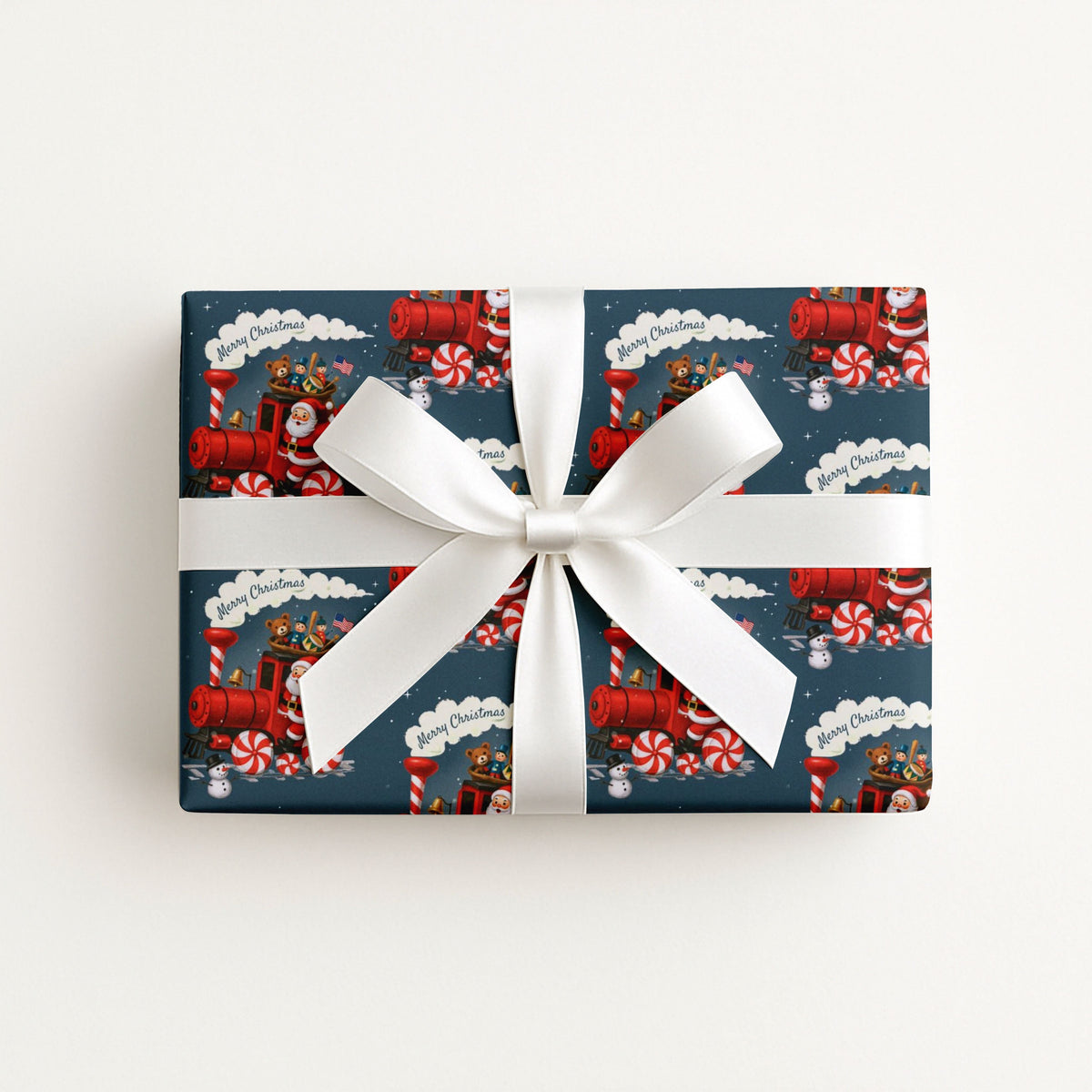 A rectangular gift box wrapped in navy holiday-themed wrapping paper with a red Santa Claus train pattern, tied with a wide white satin ribbon and bow.