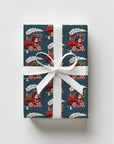 A rectangular gift box wrapped in dark blue holiday paper with a red Santa train and candy cane pattern, tied with a white ribbon and bow.