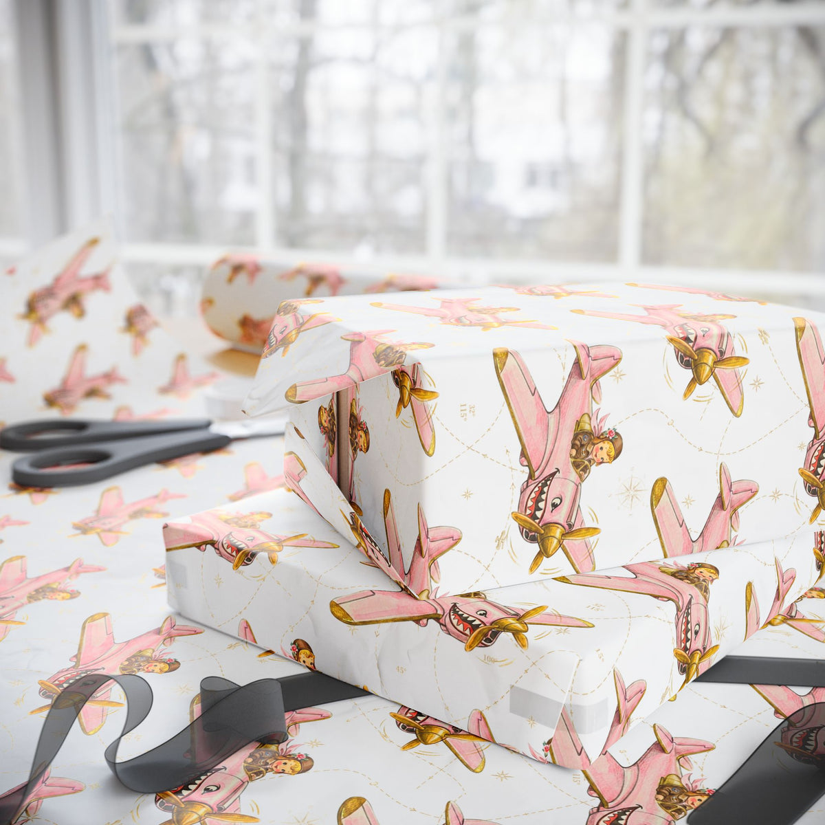 Ivory wrapping paper with a repeating pattern of vintage pink and gold airplanes, shown wrapped around gift boxes on a table with scissors and ribbon nearby.