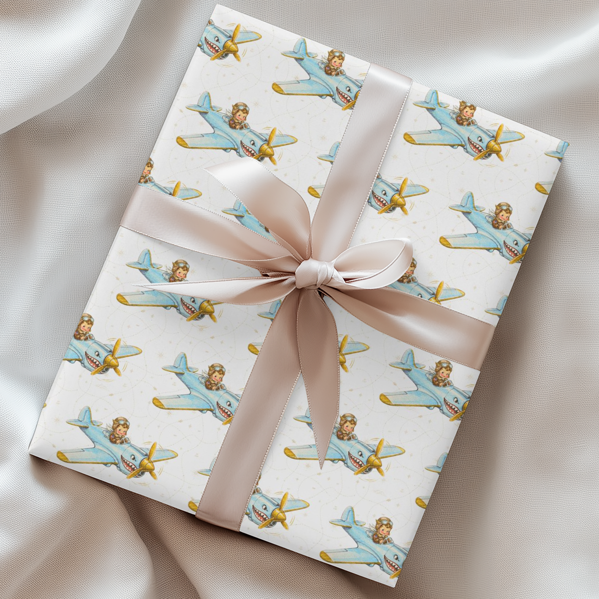 A gift box wrapped in ivory paper with a repeating pattern of light blue vintage airplanes and gold accents, tied with a wide cream satin ribbon and bow, set on a draped cream fabric background.