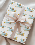 A gift box wrapped in ivory paper with a repeating pattern of light blue vintage airplanes and gold accents, tied with a wide cream satin ribbon and bow, set on a draped cream fabric background.