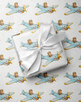 Wrapped gift tied with a white satin bow, displayed on matching wrapping paper featuring a little pilot flying a blue warplane with a painted shark mouth over a delicate navigation map with gold accents on an ivory background.