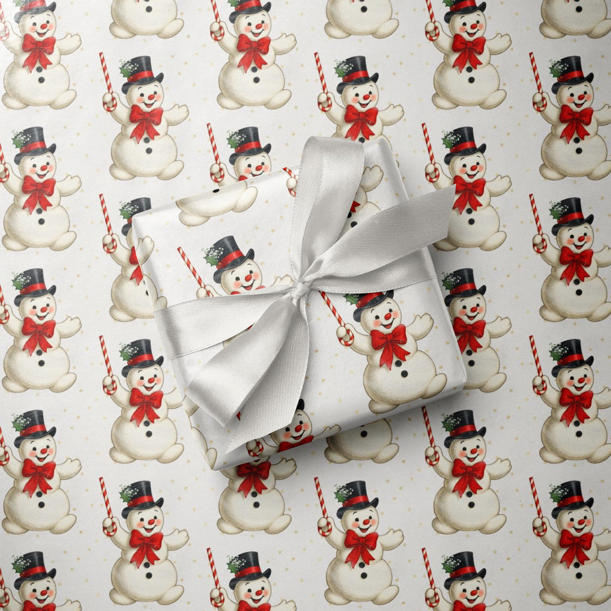 A gift box wrapped in ivory paper with a large white satin bow, placed on top of wrapping paper printed with a repeating Christmas snowman pattern.