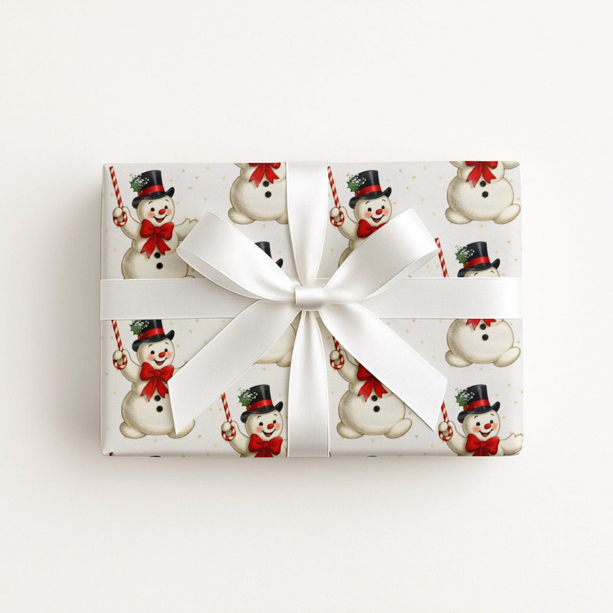 A rectangular gift box wrapped in ivory Christmas paper with a repeating snowman pattern, tied with a wide white satin ribbon and bow, photographed from above on a plain white background.