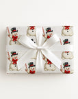 A rectangular gift box wrapped in ivory Christmas paper with a repeating snowman pattern, tied with a wide white satin ribbon and bow, photographed from above on a plain white background.