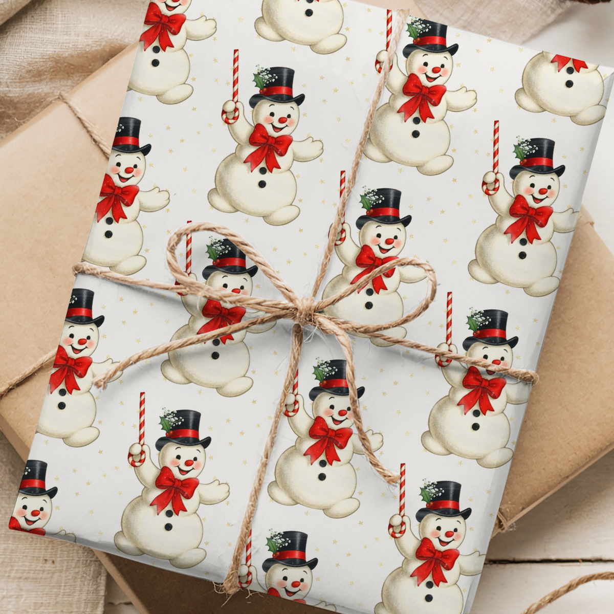 A gift-wrapped package wrapped in ivory holiday paper with a repeating snowman pattern, tied with a twine bow, placed on top of kraft paper envelopes.