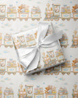 A gift box wrapped in matching baby-themed train patterned wrapping paper, tied with a white satin ribbon, placed on top of the same patterned wrapping paper.