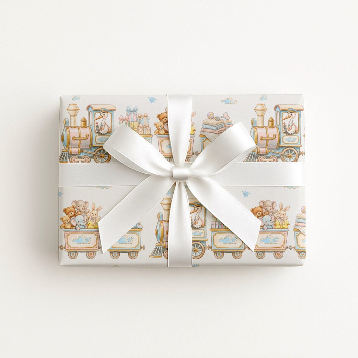 A rectangular gift box wrapped in ivory wrapping paper with a whimsical stork and train illustration pattern and tied with a wide white satin ribbon bow.