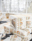 A stack of gift boxes wrapped in ivory wrapping paper with a vintage illustrated pattern of storks and trains, set on a table with scissors and ribbon.