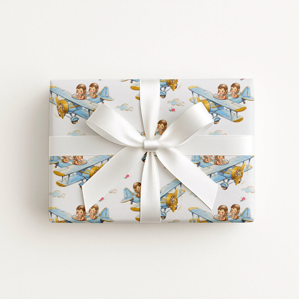 A rectangular gift box wrapped in white paper with a repeating print of vintage blue airplanes carrying twin boys, tied with a wide white satin ribbon and bow, on a plain white background.