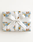 A rectangular gift box wrapped in white paper with a repeating print of vintage blue airplanes carrying twin boys, tied with a wide white satin ribbon and bow, on a plain white background.