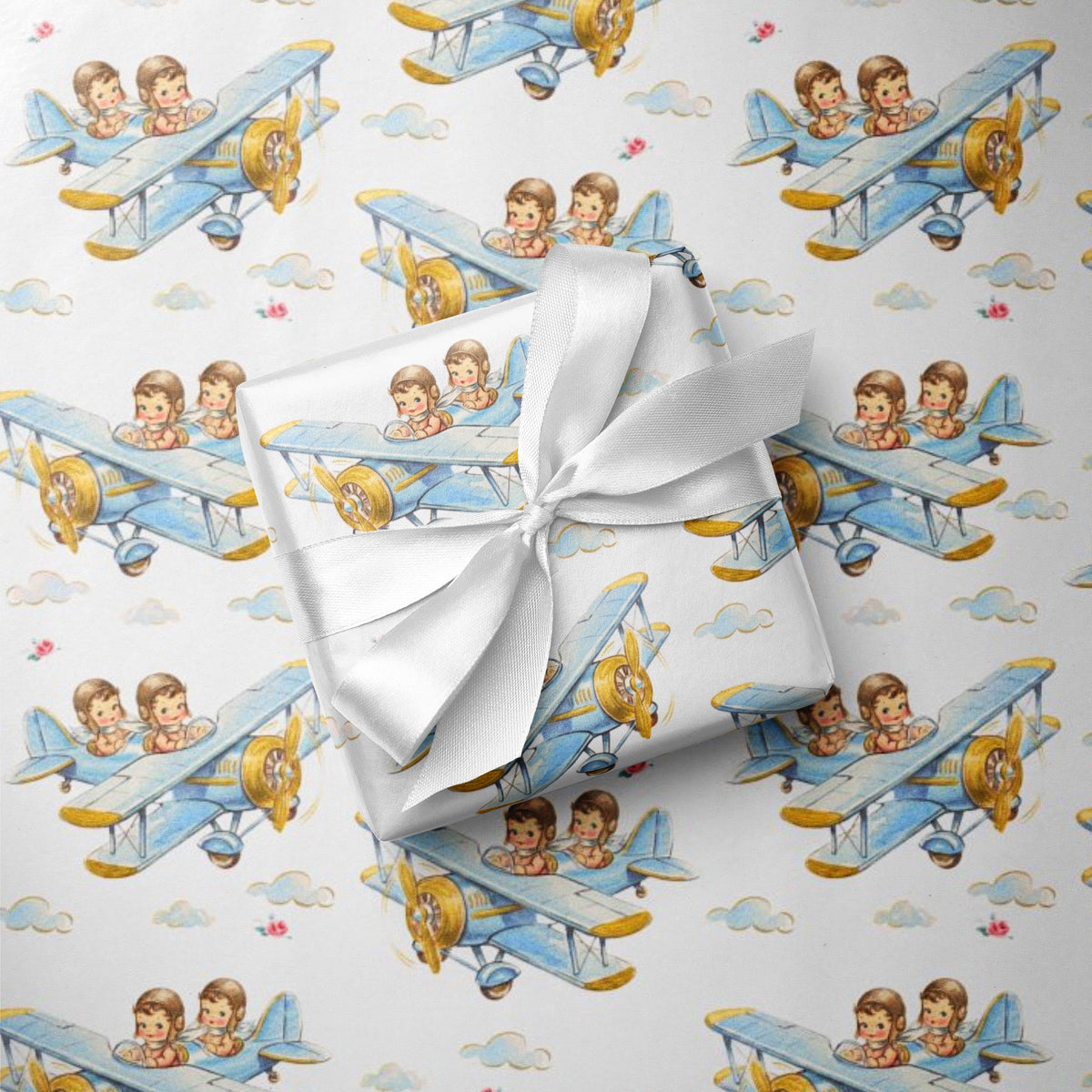 A gift box wrapped in white satin ribbon, placed on top of wrapping paper printed with a repeating pattern of blue biplanes piloted by twin boys