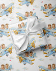 A gift box wrapped in white satin ribbon, placed on top of wrapping paper printed with a repeating pattern of blue biplanes piloted by twin boys