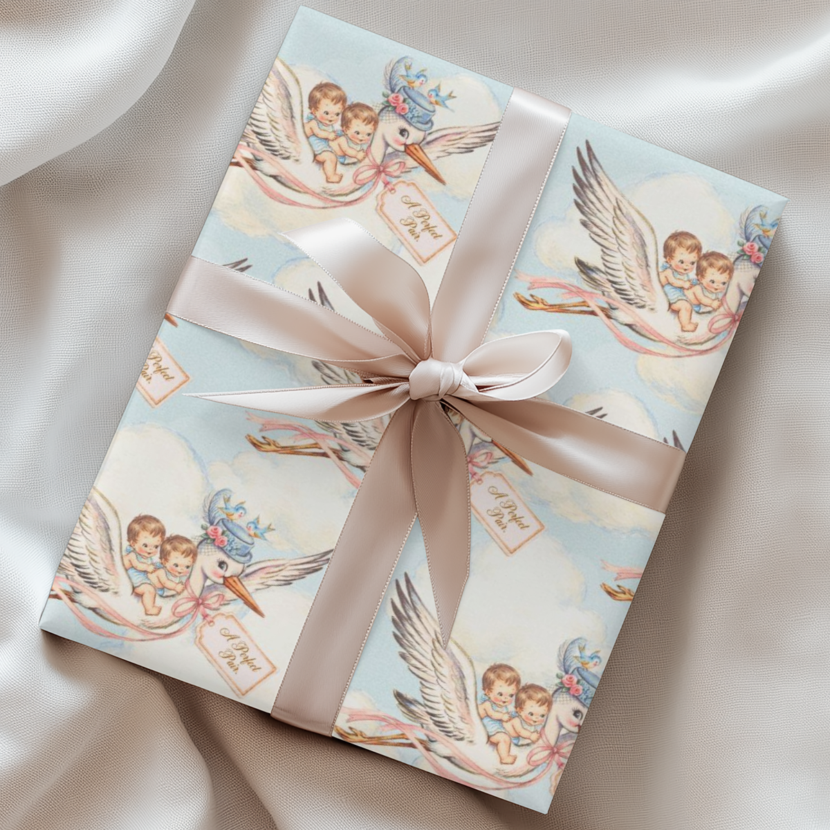 Twin boys stork wrapping paper on a gift with a blush ribbon resting on soft silk