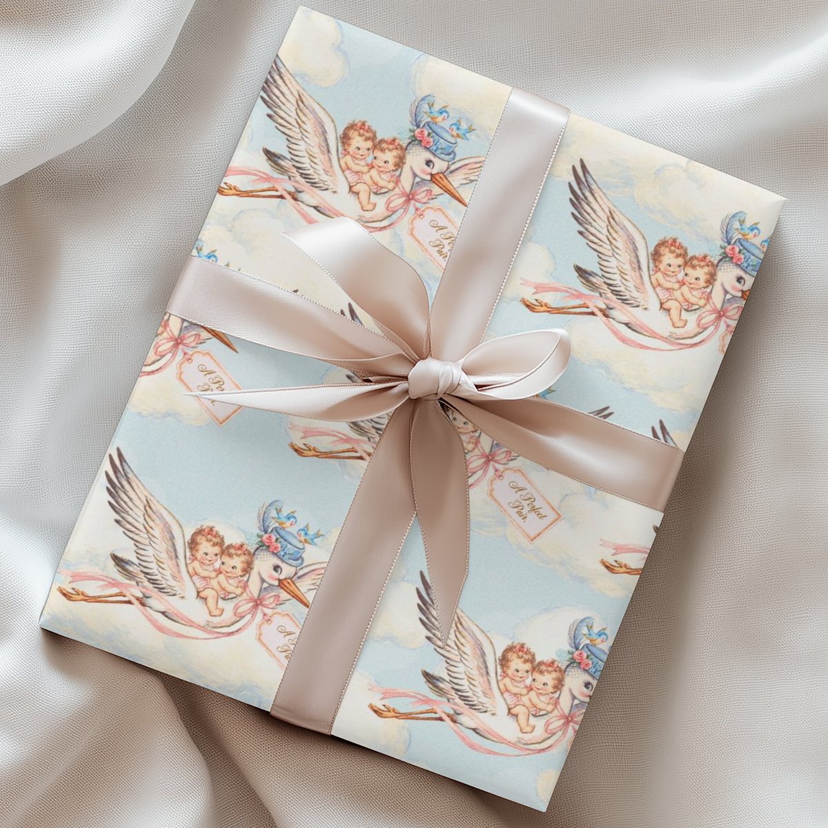 Twin girls stork wrapping paper on a gift with a blush ribbon resting on soft silk
