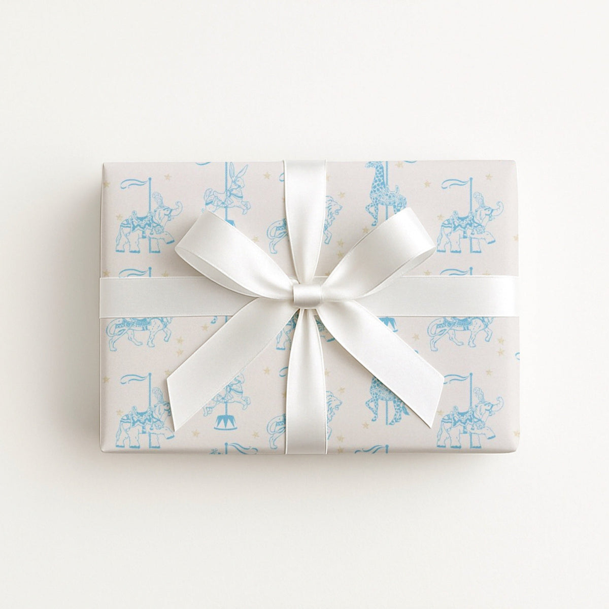 A rectangular gift box wrapped in ivory paper with a light blue carousel pattern and tied with a wide white satin ribbon bow, shown from above on a plain white background.