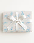 A rectangular gift box wrapped in ivory paper with a light blue carousel pattern and tied with a wide white satin ribbon bow, shown from above on a plain white background.