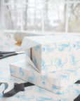 Ivory wrapping paper with a light blue carousel animal pattern, shown wrapped around stacked gift boxes on a table with scissors and ribbon near a bright window.