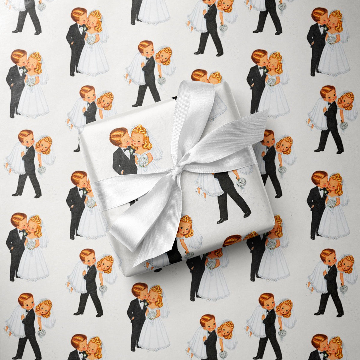 A gift box wrapped in white paper with a large white satin bow, placed on top of wedding-themed wrapping paper featuring midcentury illustrated bride and groom in black tuxedos and white wedding dresses.