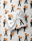 A gift box wrapped in white paper with a large white satin bow, placed on top of wedding-themed wrapping paper featuring midcentury illustrated bride and groom in black tuxedos and white wedding dresses.