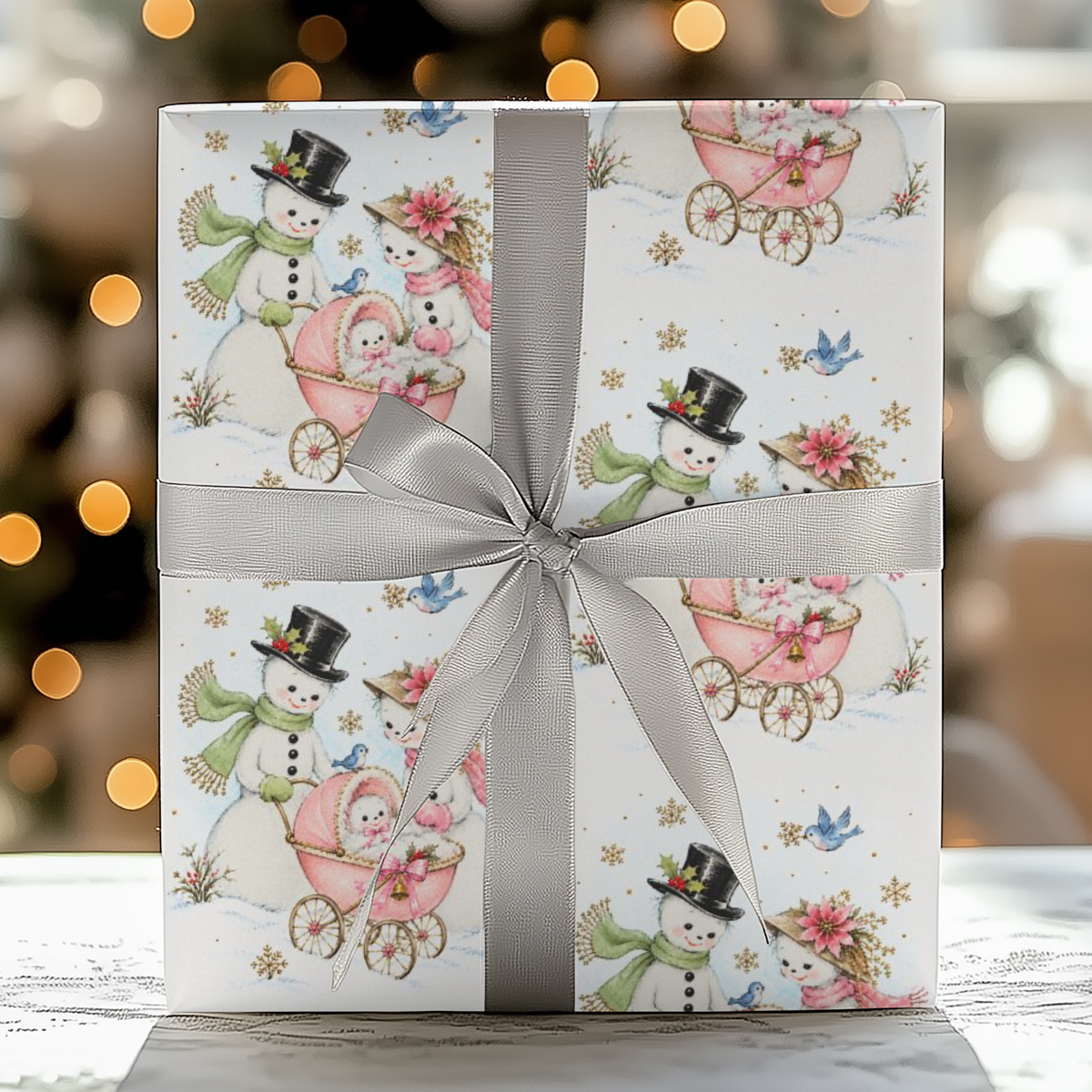 Gift wrapped in vintage-style snowman baby girl Christmas wrapping paper with a pink baby carriage and a gray satin ribbon, glowing holiday lights behind