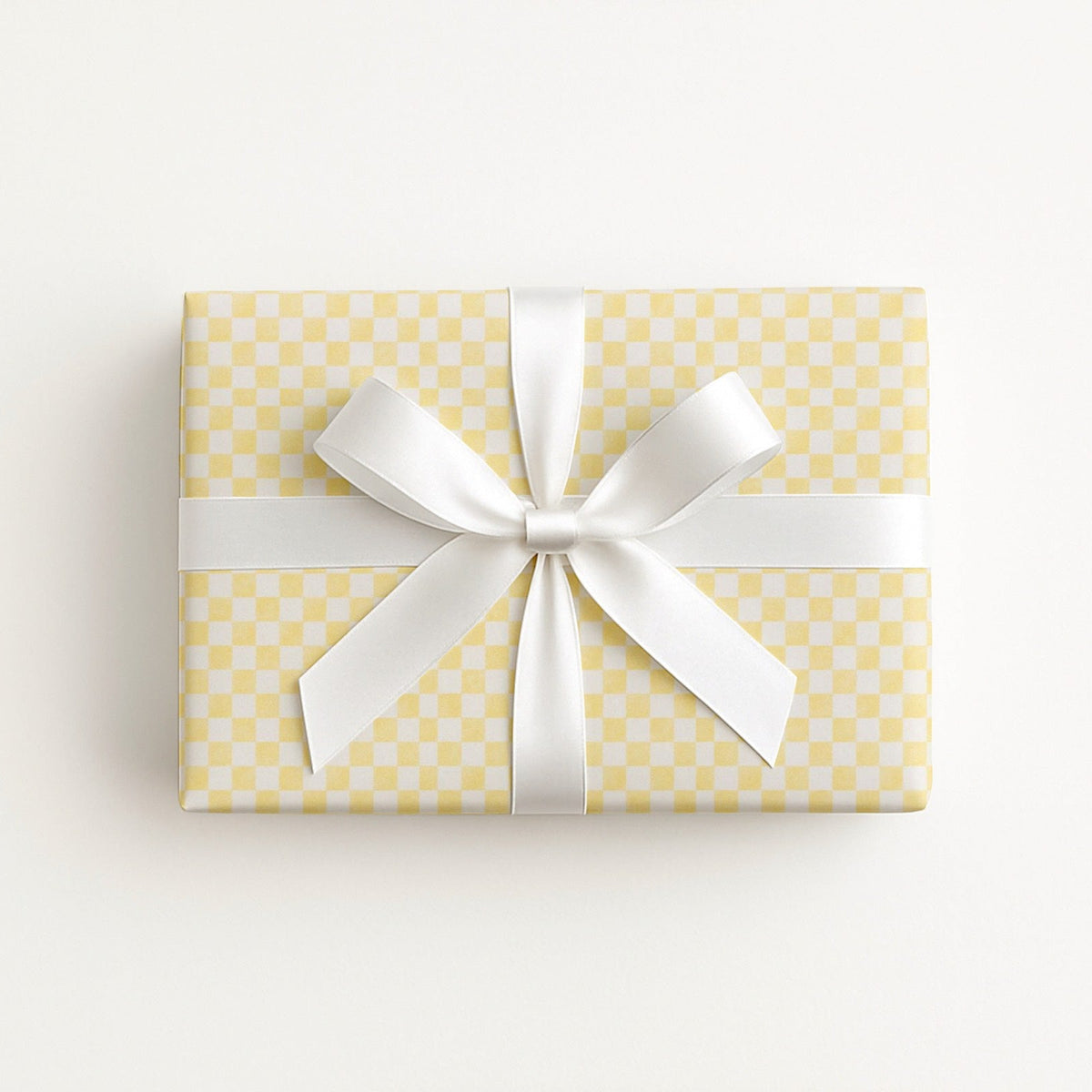 A rectangular gift box wrapped in pale yellow and white checkered paper with a wide white satin ribbon tied in a bow, photographed from above on a clean white background.