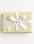 A rectangular gift box wrapped in pale yellow and white checkered paper with a wide white satin ribbon tied in a bow, photographed from above on a clean white background.