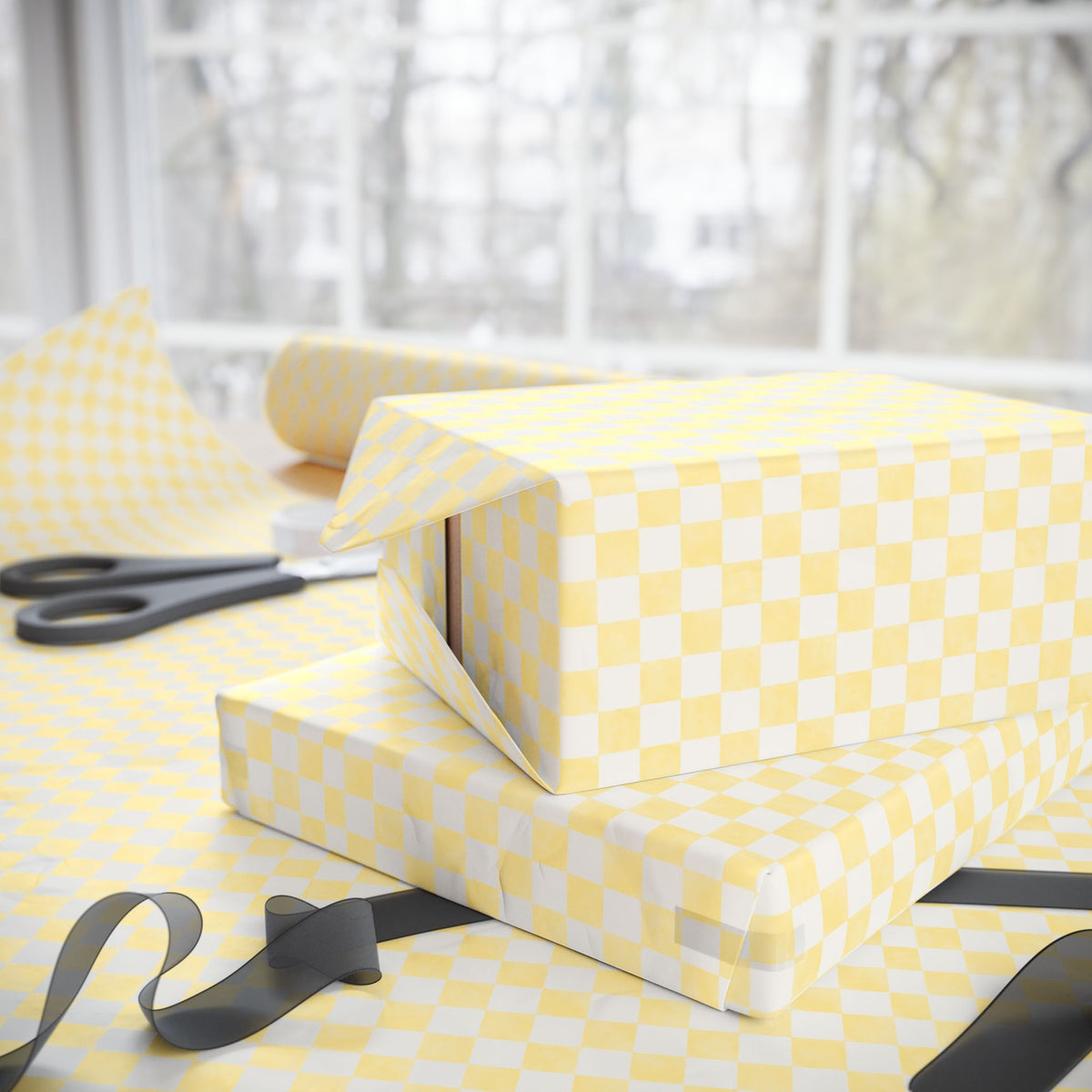 Yellow and soft white checkered wrapping paper wrapped around two gift boxes on a table, with scissors and ribbon nearby in front of a softly blurred window background.