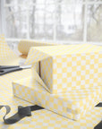 Yellow and soft white checkered wrapping paper wrapped around two gift boxes on a table, with scissors and ribbon nearby in front of a softly blurred window background.